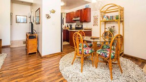 A dining area with a round table and wicker chairs, a kitchen in the background, and a bookshelf with books and decorative items. - Island Touches w/Modern Kitchen, Wood Floors, Lanai, Flat Screen, WiFi–Kaha Lani 203 (LIHUE)
