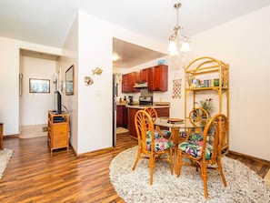 A dining area with a round table and wicker chairs, a kitchen in the background, and a bookshelf with books and decorative items. - Island Touches w/Modern Kitchen, Wood Floors, Lanai, Flat Screen, WiFi–Kaha Lani 203 (LIHUE)