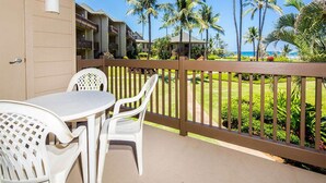 A balcony with a table and chairs, overlooking a pool and palm trees, with buildings in the background. - Island Touches w/Modern Kitchen, Wood Floors, Lanai, Flat Screen, WiFi–Kaha Lani 203 (LIHUE)