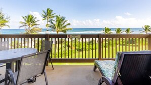 A covered outdoor area with a table and chairs, a sign indicating a restaurant, and a view of the ocean and palm trees. - 1 Bedroom Condo w/Epic Ocean View - Kaha Lani 319 (LIHUE)
