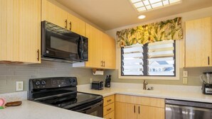 A modern kitchen with wooden cabinets, a black microwave, a stove, a dishwasher, and a window with a tropical view. - 1 Bedroom Condo w/Epic Ocean View - Kaha Lani 319 (LIHUE)
