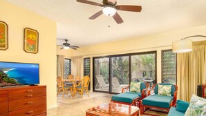 A spacious living room with a television, wooden furniture, a ceiling fan, and large windows offering a view of the outdoors.