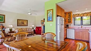 A dining area with a wooden table and chairs, a kitchen with wooden cabinets, and a living room with a sofa and a red chair. - Ground-Floor Ocean View W/Lanai–Kaha Lani #105 (LIHUE)