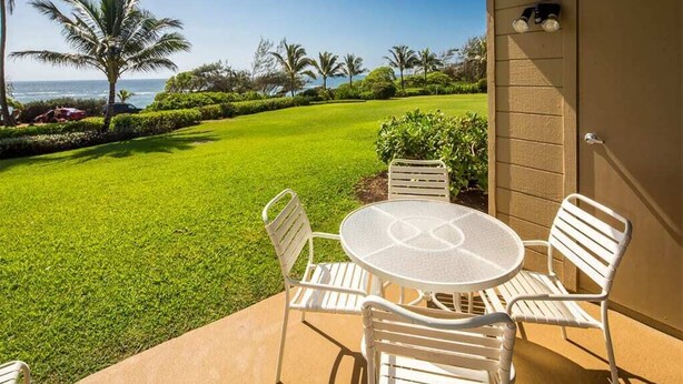 A patio with a round table and white chairs, overlooking a well-manicured lawn and palm trees, with a view of the ocean. - Ocean Suite Life Steps from Surf – Kaha Lani 120 (LIHUE)