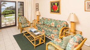 A living room with a sofa, two armchairs, a coffee table, and a floor lamp. There is a sliding glass door leading to a patio with outdoor furniture. - Ocean Suite Life Steps from Surf – Kaha Lani 120 (LIHUE)