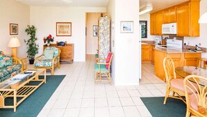 A spacious living room with a sofa, armchairs, and a coffee table. A kitchen with wooden cabinets and a microwave is visible in the background. - Ocean Suite Life Steps from Surf – Kaha Lani 120 (LIHUE)