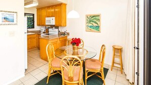 A compact kitchen with wooden cabinets, a round glass dining table, and wicker chairs. - Ocean Suite Life Steps from Surf – Kaha Lani 120 (LIHUE)