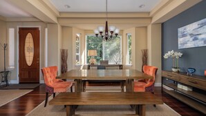 A dining area with a wooden table, two orange chairs, a chandelier, and a view of greenery through large windows. - Sierra Sky - Luxurious Retreat with Free Private Lake Access! (Arnold)