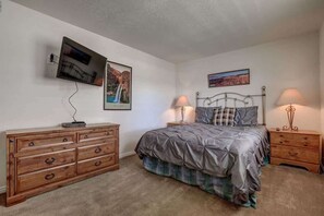 A bedroom with a bed, two bedside tables with lamps, a dresser, a TV mounted on the wall, and two framed pictures. - Las Palmas Resort #205 | All-Inclusive Amenities (Saint George)