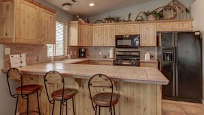 A kitchen with wooden cabinets, a central island with bar stools, a refrigerator, and a microwave. - Las Palmas Resort #1018 | All-Inclusive Amenities (Saint George)