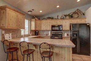 A kitchen with wooden cabinets, a central island with bar stools, a refrigerator, and a microwave. - Las Palmas Resort #1018 | All-Inclusive Amenities (Saint George)
