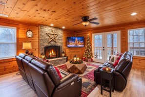 Interior acogedor de una cabaña con chimenea de piedra, sofás de cuero, un árbol de Navidad y un televisor.