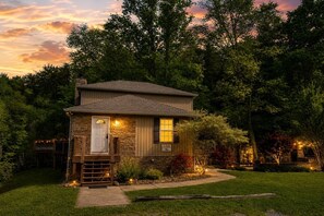A small stone house with a front porch, a wooden deck, and a well-lit entrance.