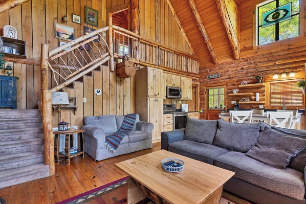 A rustic cabin interior with a wooden staircase, two sofas, a wooden coffee table, and a kitchen area.