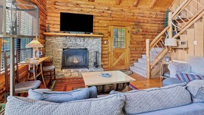 Ein Blockhaus-Interieur mit Kamin aus Stein, Holzwänden, einem über dem Kamin montierten Fernseher und einer Treppe, die zu einer oberen Ebene führt.