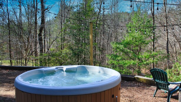 A hot tub surrounded by a forested area with a chair and a table nearby. - HotTub, OutdoorShower, Renovated Barndominium, WiFi, Fireplace, Pet Friendly (Ferguson)