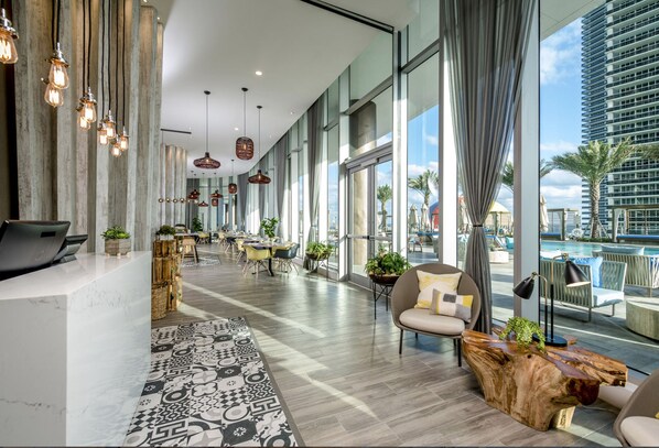 A modern hotel lobby with a long reception desk, seating area, and large windows offering views of a pool and cityscape.