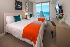 Habitación de hotel con dos camas, un escritorio, una silla, un televisor y un ventanal con vista a la ciudad y al océano.