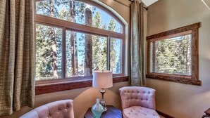 Une pièce comprenant deux fauteuils roses à motifs capitonnés, une petite table ronde avec une lampe et un vase, et deux fenêtres donnant sur des arbres.