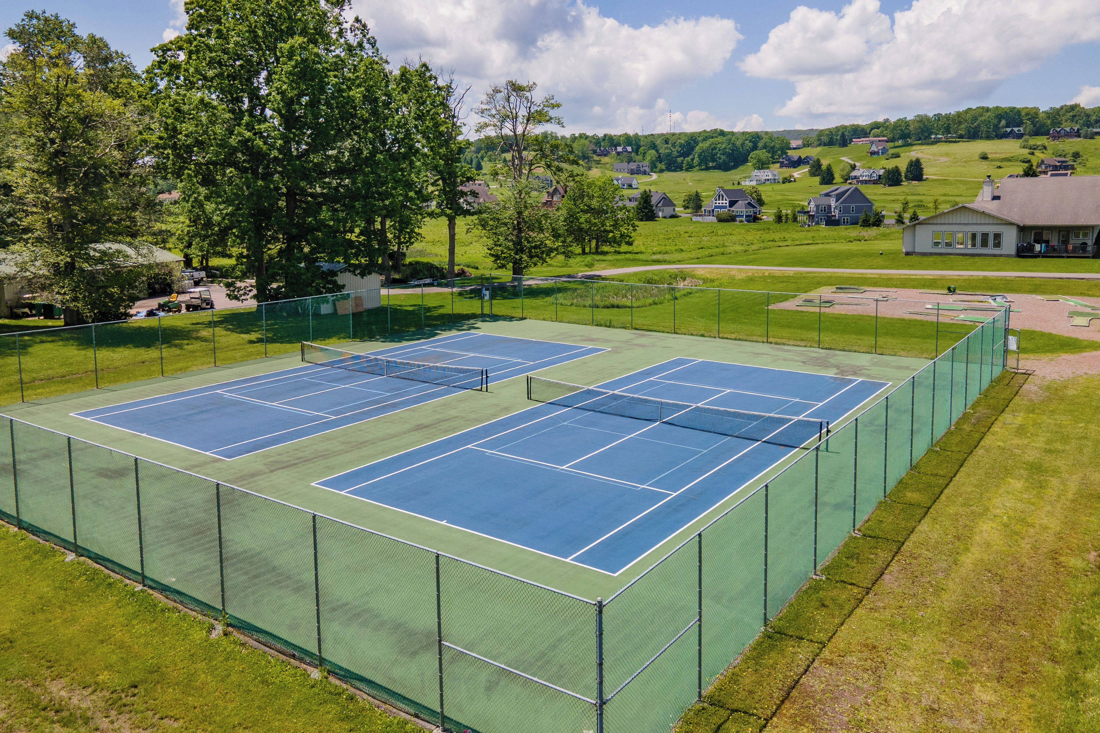 A tennis court with a net, surrounded by a green fence, set against a backdrop of houses and trees.