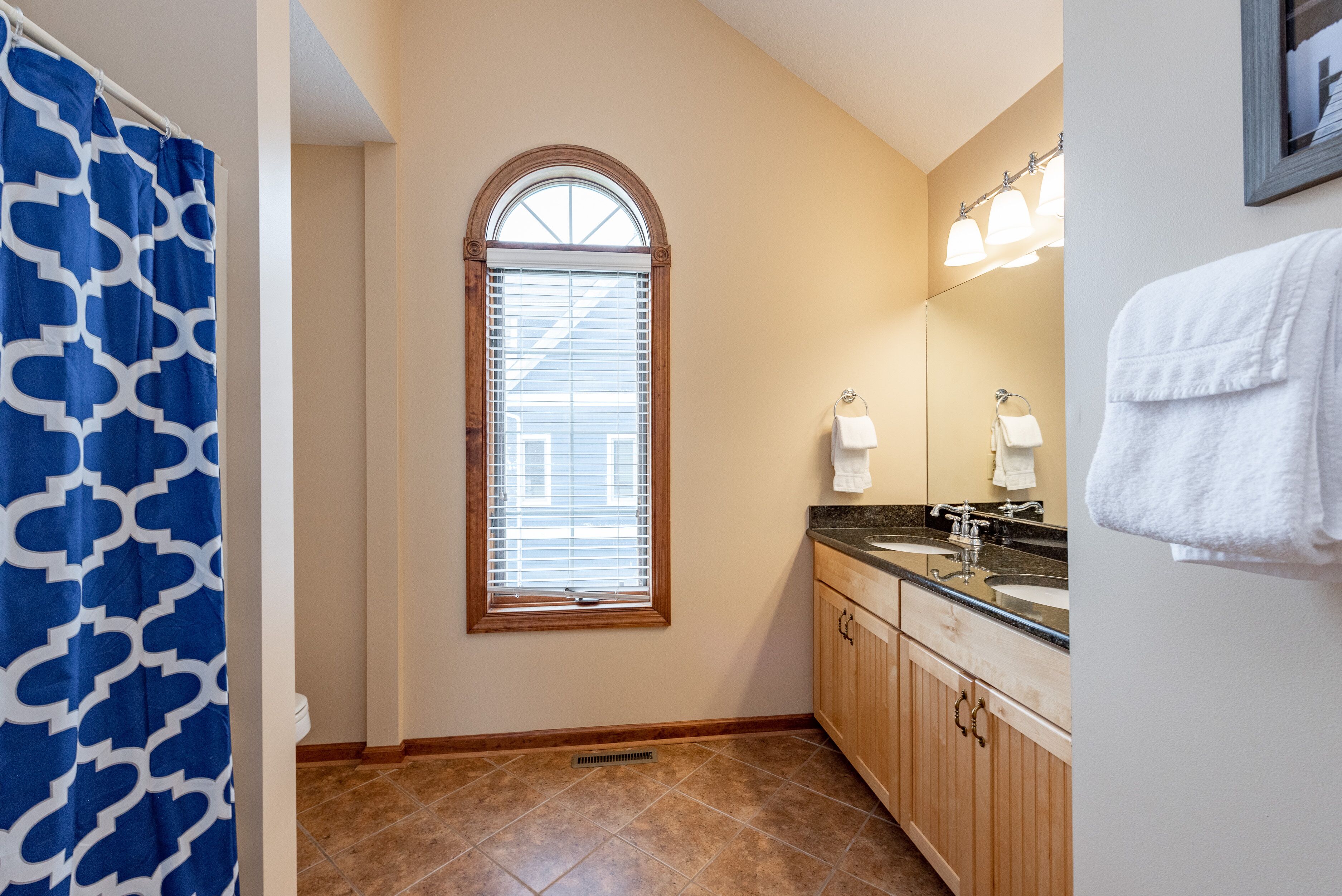 A bathroom with a shower curtain, a window with blinds, wooden cabinets, a sink with a mirror, and a towel hanging on the wall.