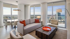 Ein modernes Wohnzimmer mit einem grauen Sofa, beerenfarbenen Sesseln, einem runden Couchtisch und einem Esstisch mit Stühlen. Große Fenster bieten Blick auf die Stadt und den Ozean.