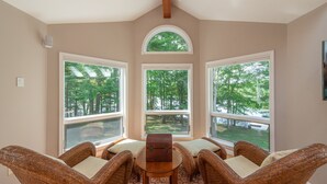 A room with a large window, wicker furniture, and a wooden table. - Luxury Lakefront Gem w/ Hot Tub & Serenity (Oakland)