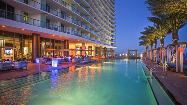 Zona de piscina de hotel con varios niveles de balcones, mobiliario exterior y vista despejada al agua.