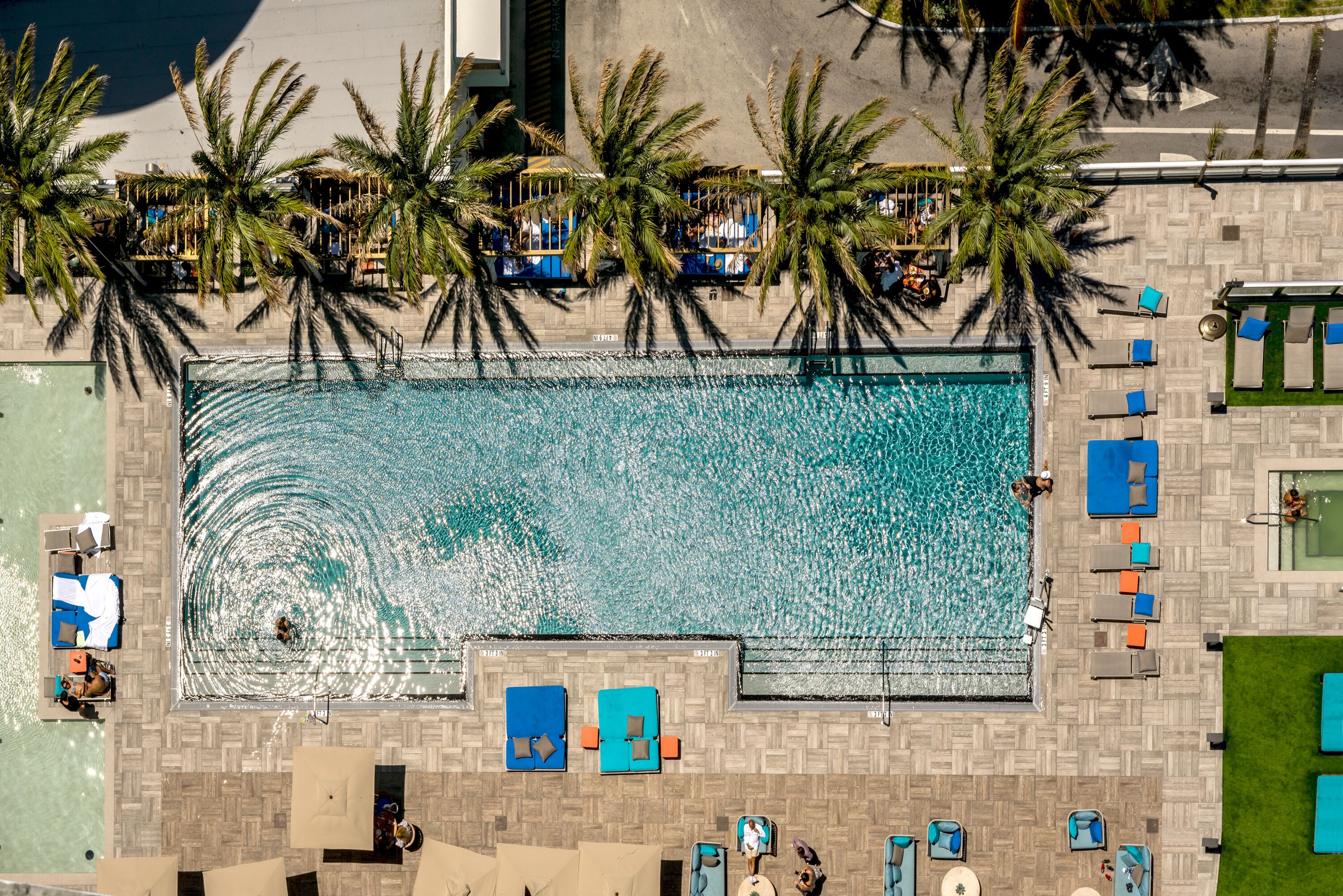 Eine Vogelperspektive zeigt ein Resort mit großem Schwimmbecken, Palmen und Liegebereichen.