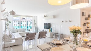 A modern living room with a sofa, clear chairs, a dining table set for six, and a large window with a city view. - Cozy designer condo across from the beach. WiFi, parking, tennis and more! (Sunny Isles Beach.)