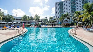 A swimming pool with lounge chairs and umbrellas, surrounded by palm trees and a tall building in the background. - Cozy designer condo across from the beach. WiFi, parking, tennis and more! (Sunny Isles Beach.)