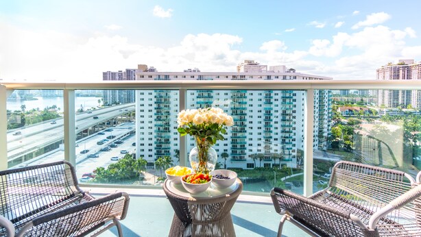 A balcony with wicker chairs, a table with a vase of flowers, and a view of a cityscape with high-rise buildings and a highway. - Cozy designer condo across from the beach. WiFi, parking, tennis and more! (Sunny Isles Beach.)