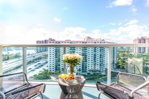 A balcony with wicker chairs, a table with a vase of flowers, and a view of a cityscape with high-rise buildings and a highway. - Cozy designer condo across from the beach. WiFi, parking, tennis and more! (Sunny Isles Beach.)