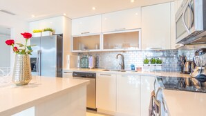 A modern kitchen with white cabinetry, a stainless steel refrigerator, a dishwasher, and a microwave. There are potted plants on the countertop and a vase with flowers on the island. - Cozy designer condo across from the beach. WiFi, parking, tennis and more! (Sunny Isles Beach.)