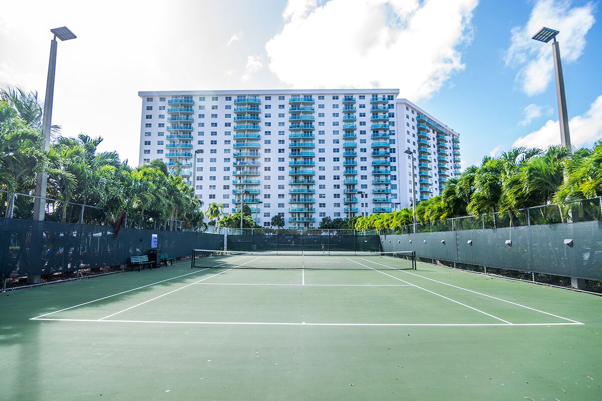 A tennis court with a net, surrounded by a fence, and a tall building in the background.