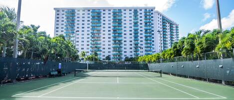 Una cancha de tenis con red, rodeada por una valla, y un edificio alto al fondo.
