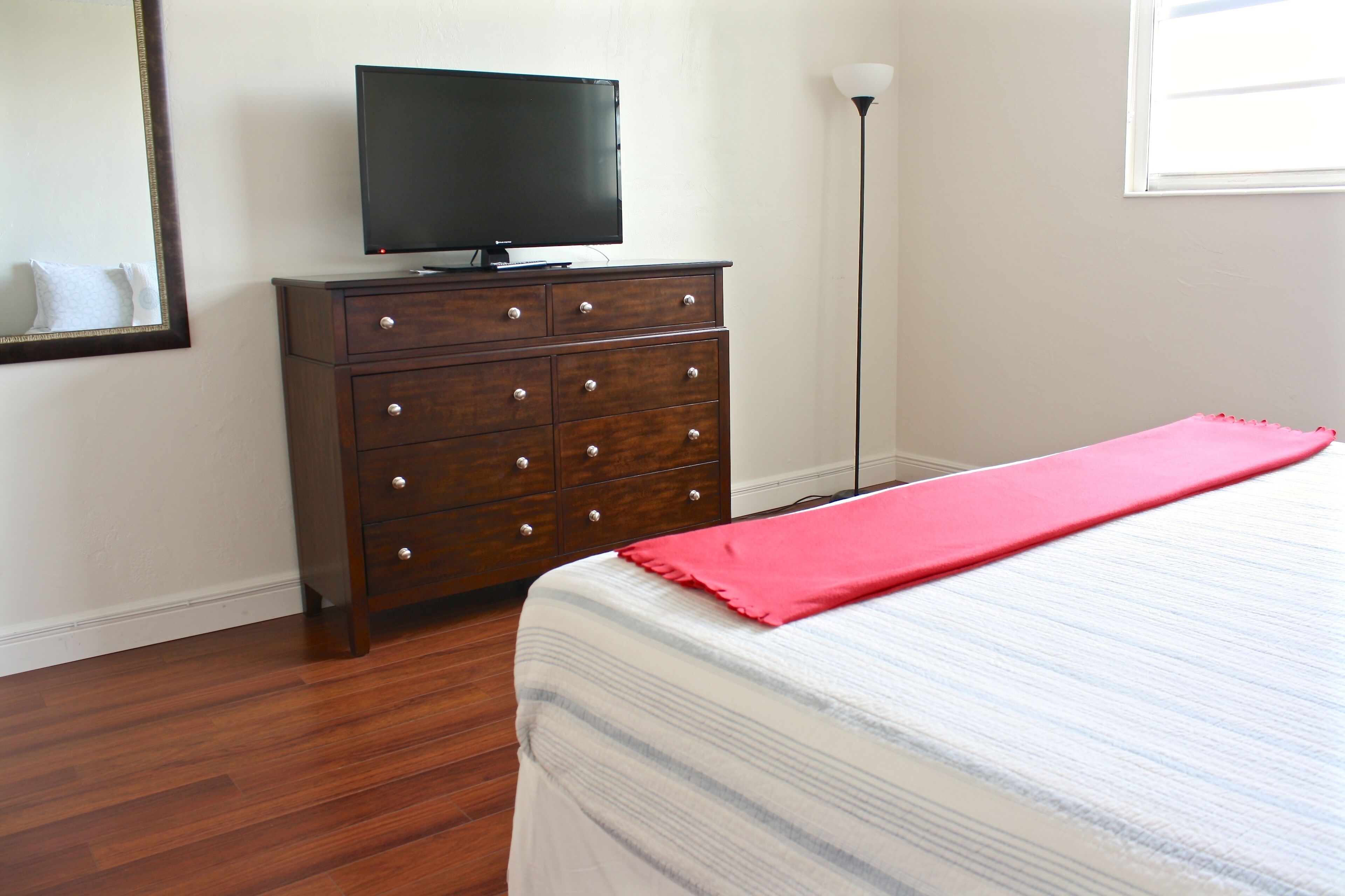 A bedroom with a wooden dresser, a television on top, a bed with a striped bedspread, and a floor lamp.