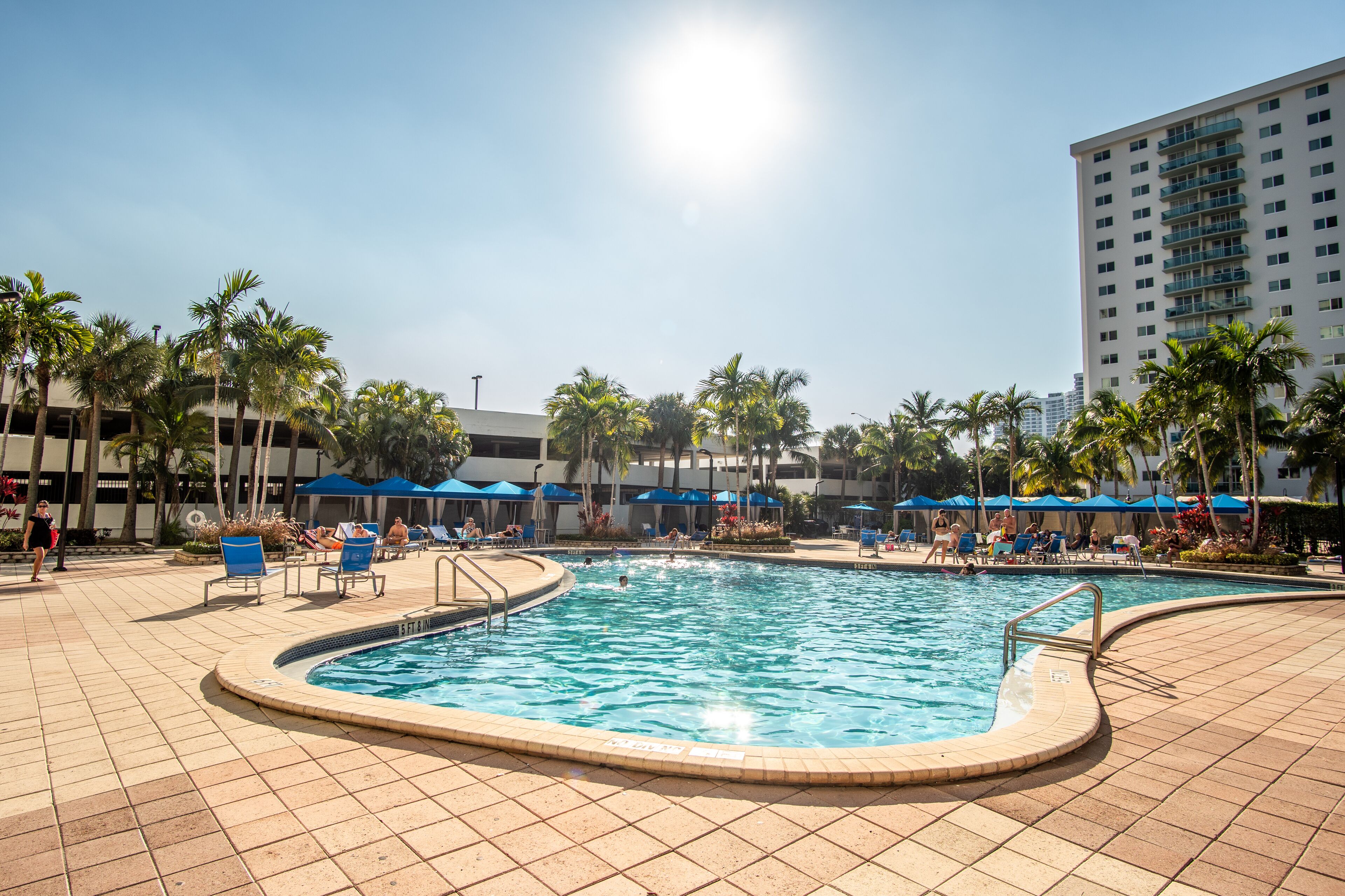 Une piscine avec une terrasse carrelée, des palmiers et des parasols bleus.