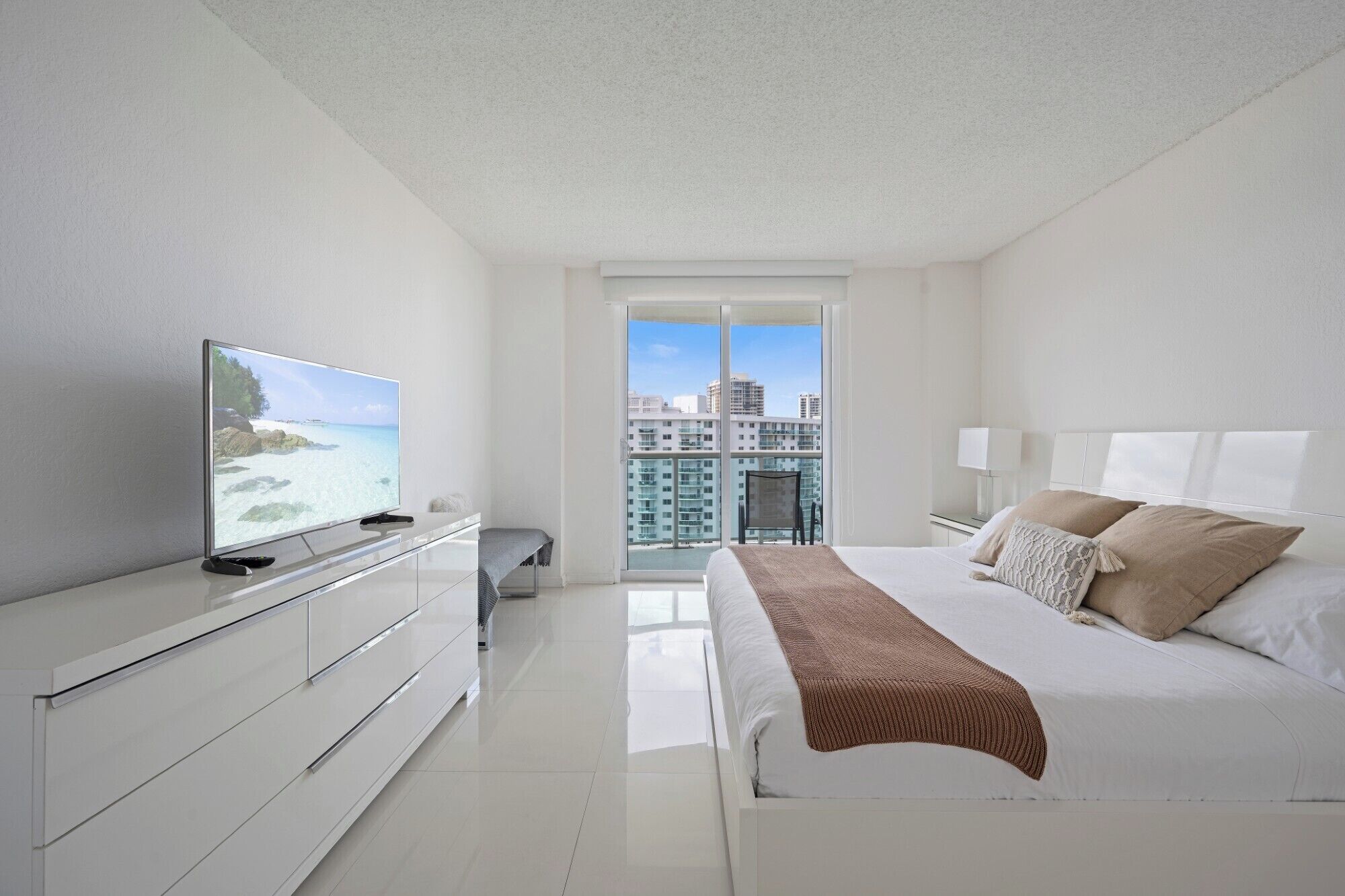 A modern hotel room with a large bed, a TV on a white dresser, and a balcony with a city view.