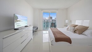 Una habitación de hotel moderna con una cama grande, un televisor sobre un tocador blanco y un balcón con vistas a la ciudad.