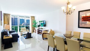A modern living room with a sofa, coffee table, dining table, and chairs. There is a TV mounted on the wall and a chandelier.
