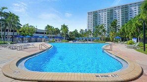 A swimming pool with lounge chairs and umbrellas, surrounded by palm trees and a tall building in the background. - Modern condo w/ bay views. WiFi, parking, tennis, playground and more! (Sunny Isles Beach.)