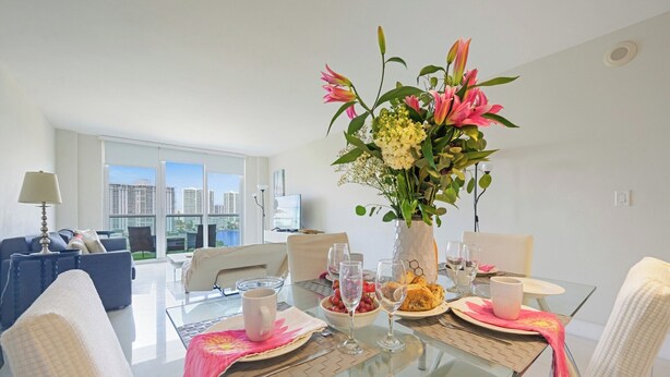 A modern dining area with a glass table, white chairs, and a vase of flowers. - Modern condo w/ bay views. WiFi, parking, tennis, playground and more! (Sunny Isles Beach.)