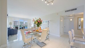 A spacious living area with a glass dining table, white chairs, and a city view through large windows. - Modern condo w/ bay views. WiFi, parking, tennis, playground and more! (Sunny Isles Beach.)