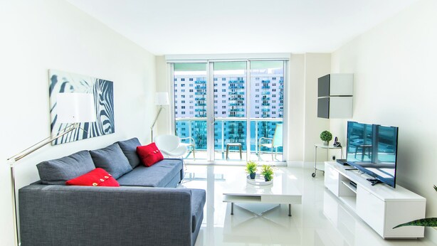 Een moderne woonkamer met een grijze bank, rode kussens, een witte salontafel, een flatscreen-tv en een groot raam met uitzicht op de stad.