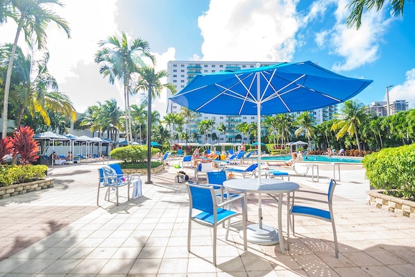 An outdoor pool area with lounge chairs and umbrellas, surrounded by palm trees and a building in the background.