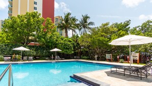 A swimming pool with lounge chairs and umbrellas, surrounded by trees and buildings. - Spacious condo w/ terrace overlooking the bay | WIFI + Parking (Sunny Isles Beach)