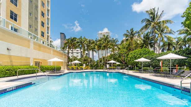 A swimming pool with lounge chairs and umbrellas, surrounded by palm trees and a building with balconies. - Spacious condo w/ terrace overlooking the bay | WIFI + Parking (Sunny Isles Beach)