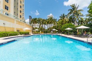 Piscina com espreguiçadeiras e guarda-sóis, cercada por palmeiras e um edifício com varandas.