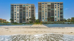 Two high-rise buildings with multiple floors, a sandy beach in the foreground, and palm trees in the background. - Reflections 804 - GULF FRONT Condo! (Indian Rocks Beach)